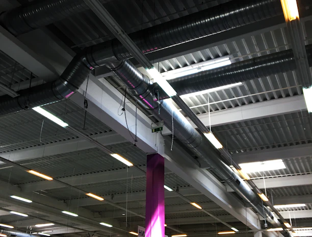 A large indoor space with an industrial-style ceiling featuring exposed ductwork and fluorescent lighting. Below, there are various goods and displays typical of a hardware or retail store with signs and product displays visible. A prominent purple column stands in the middle, surrounded by different types of merchandise.